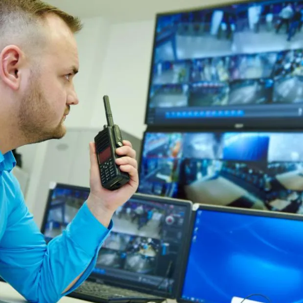 Foto ilustrativa: operador de monitoramento com rádio HT diante de uma parede de monitores com imagens de câmeras