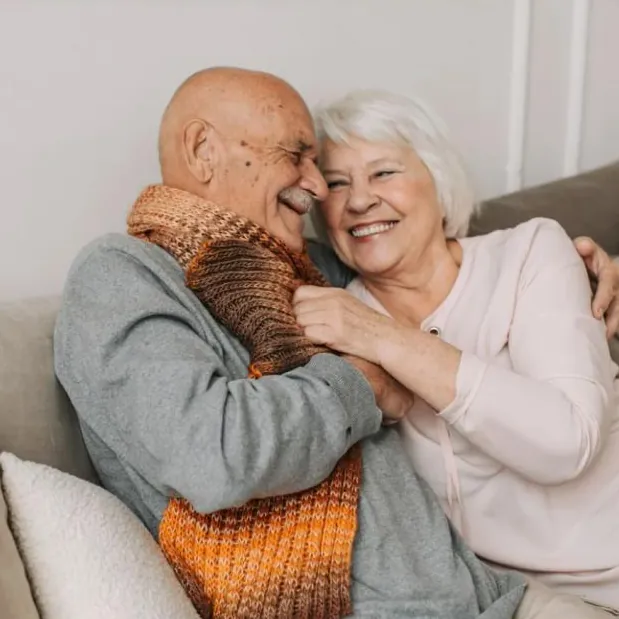 Foto ilustrativa: casal de idosos abraçado no sofá de casa